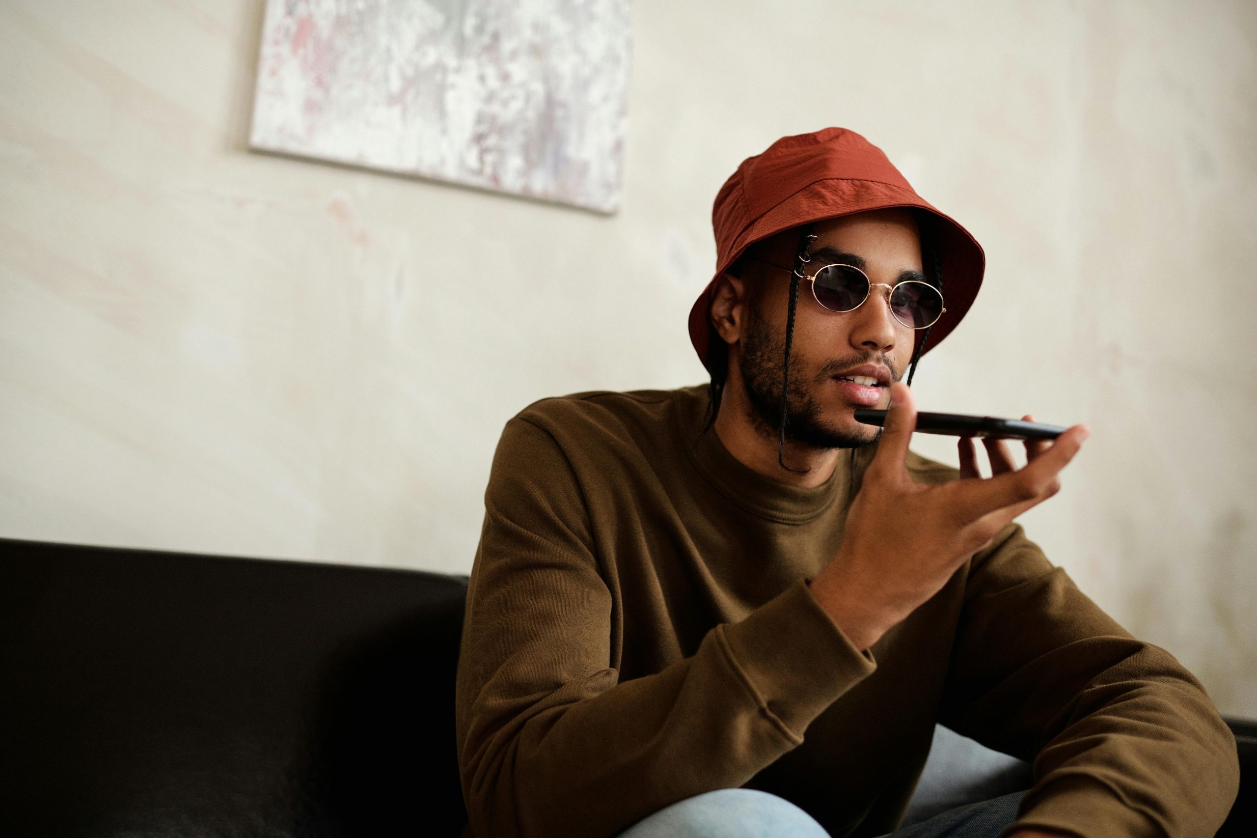 A stylish young man with sunglasses and a bucket hat using a smartphone for a voice call indoors.